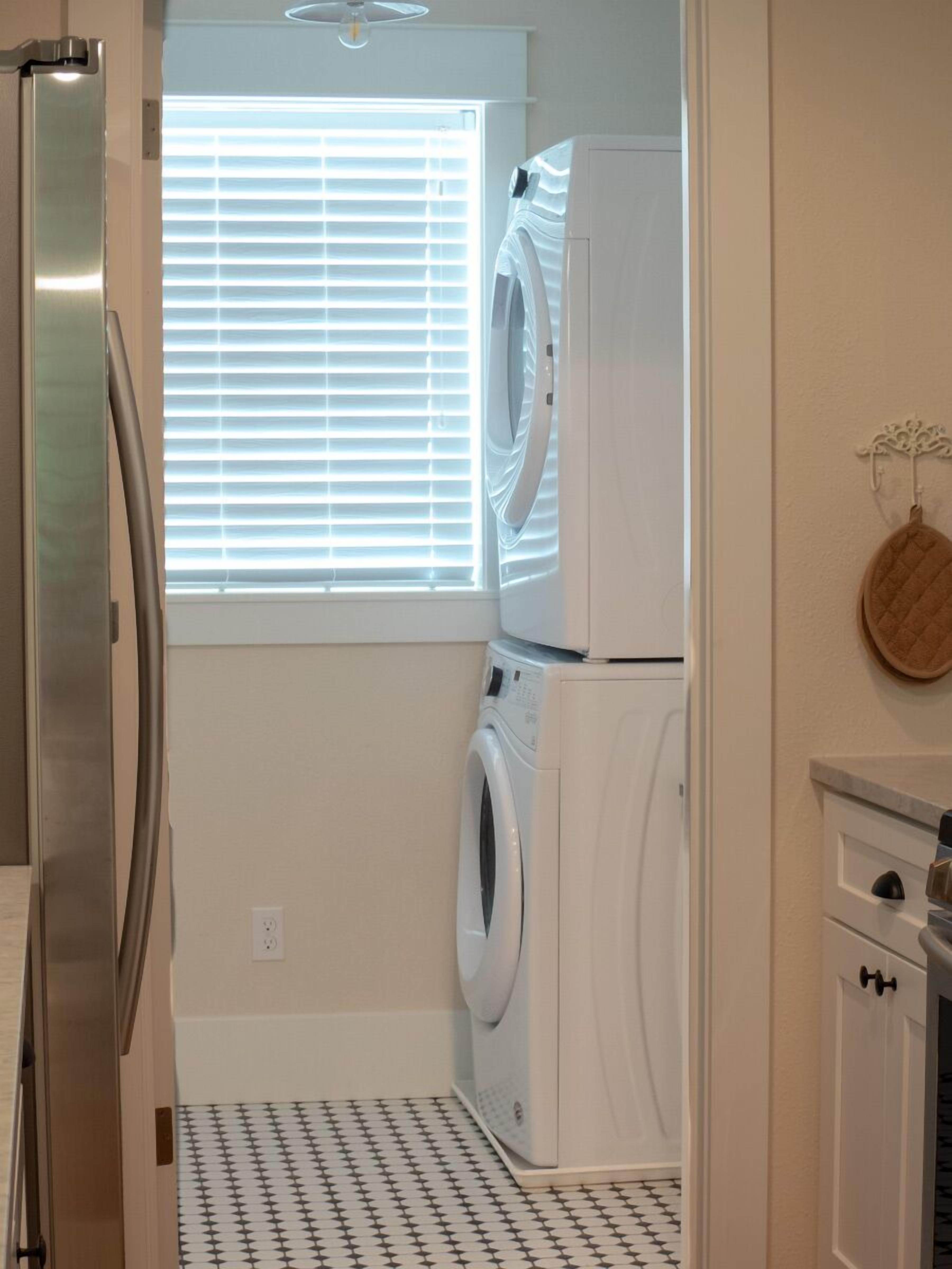 A compact laundry room with a washing machine and dryer, a stainless steel refrigerator, and a window with blinds.