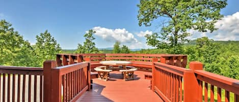 A wooden deck with a picnic table and benches, surrounded by lush greenery and a clear sky.