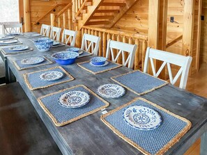 Una larga mesa de comedor con posavasos cuadrados y platos azules y blancos, rodeada de sillas blancas.