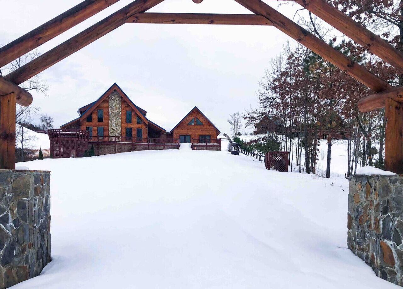 A log cabin with a stone entrance and a snow-covered driveway leading to the main building.