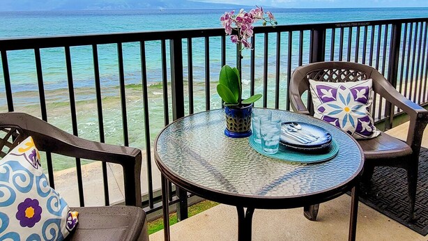 A balcony with a round glass table, two chairs, a potted plant, and a view of the ocean.