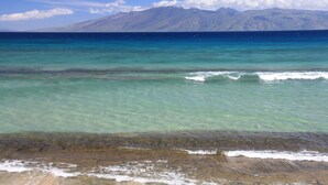 A beach with clear turquoise water, a rocky shoreline, and a mountain range in the background.