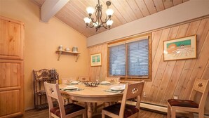 A dining area with a wooden table set for four, surrounded by wooden chairs with cushioned seats. A chandelier hangs from the ceiling, and there are two windows with blinds.
