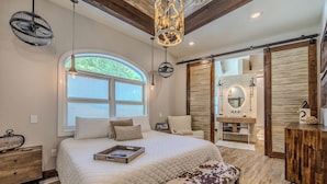 A bedroom with a large bed, a bench, a wooden dresser, a vanity, and a large window. - Upscale Lakefront Luxury at Lake of the Ozarks - Private Dock, Quiet Cove (Lake Ozark)