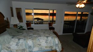 Un dormitorio con una cama grande, vistas a un atardecer sobre el agua, una silla y una lámpara.
