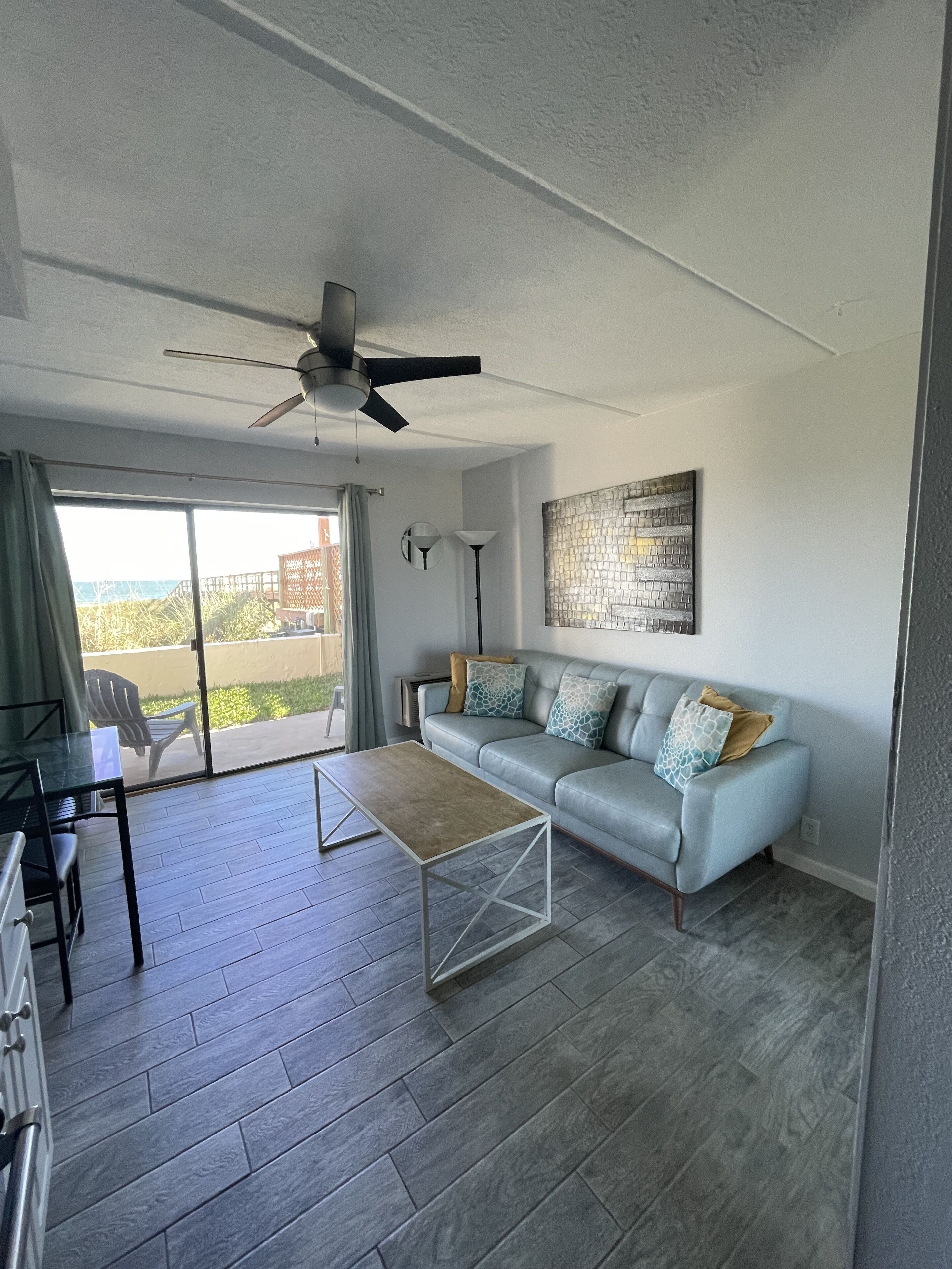A modern living room with a grey sofa, a coffee table, a ceiling fan, and a view through a sliding glass door.