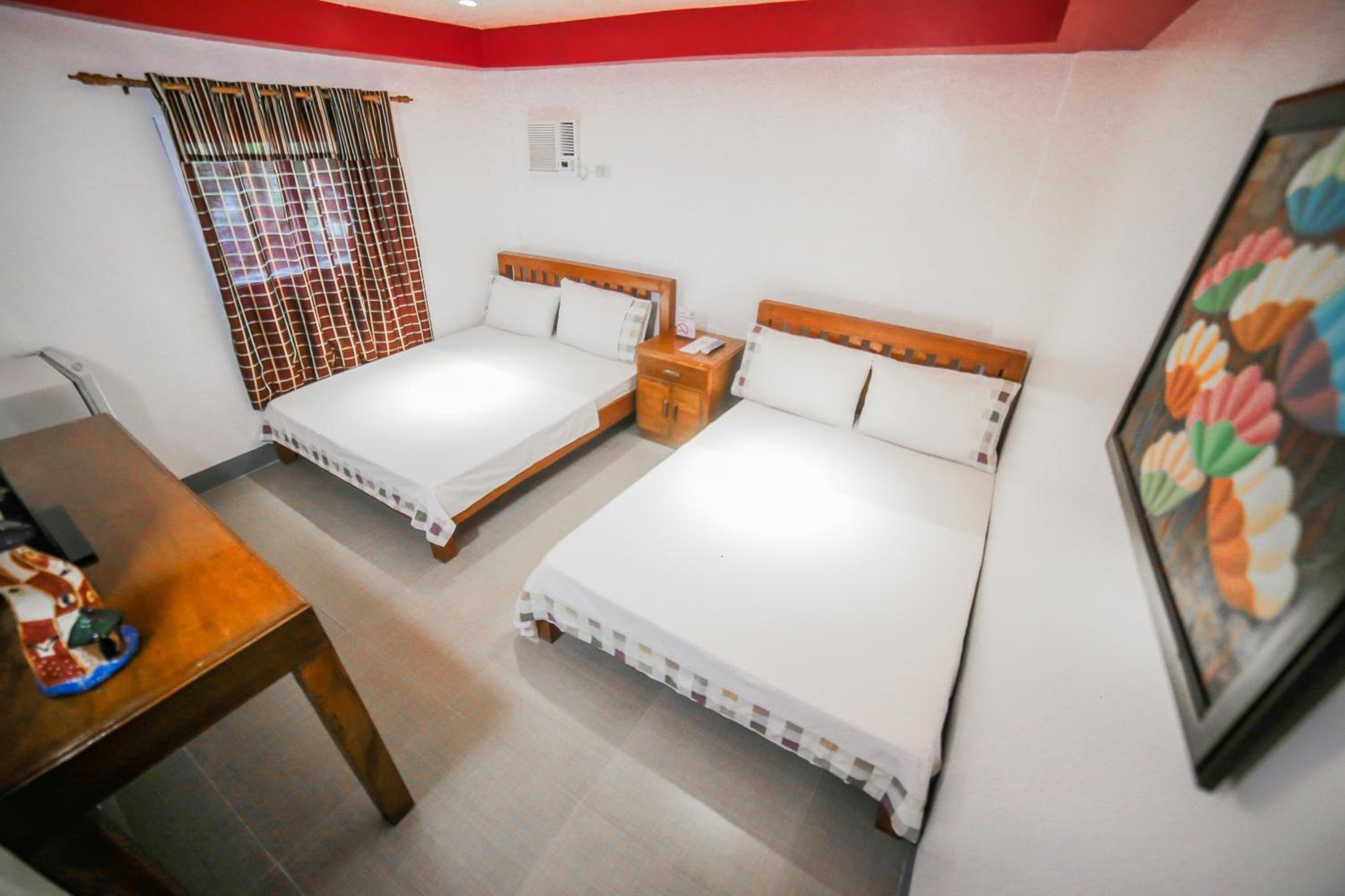 A room with two single beds, a wooden table, a small bedside table, a window with a curtain, and a wall-mounted air conditioning unit.