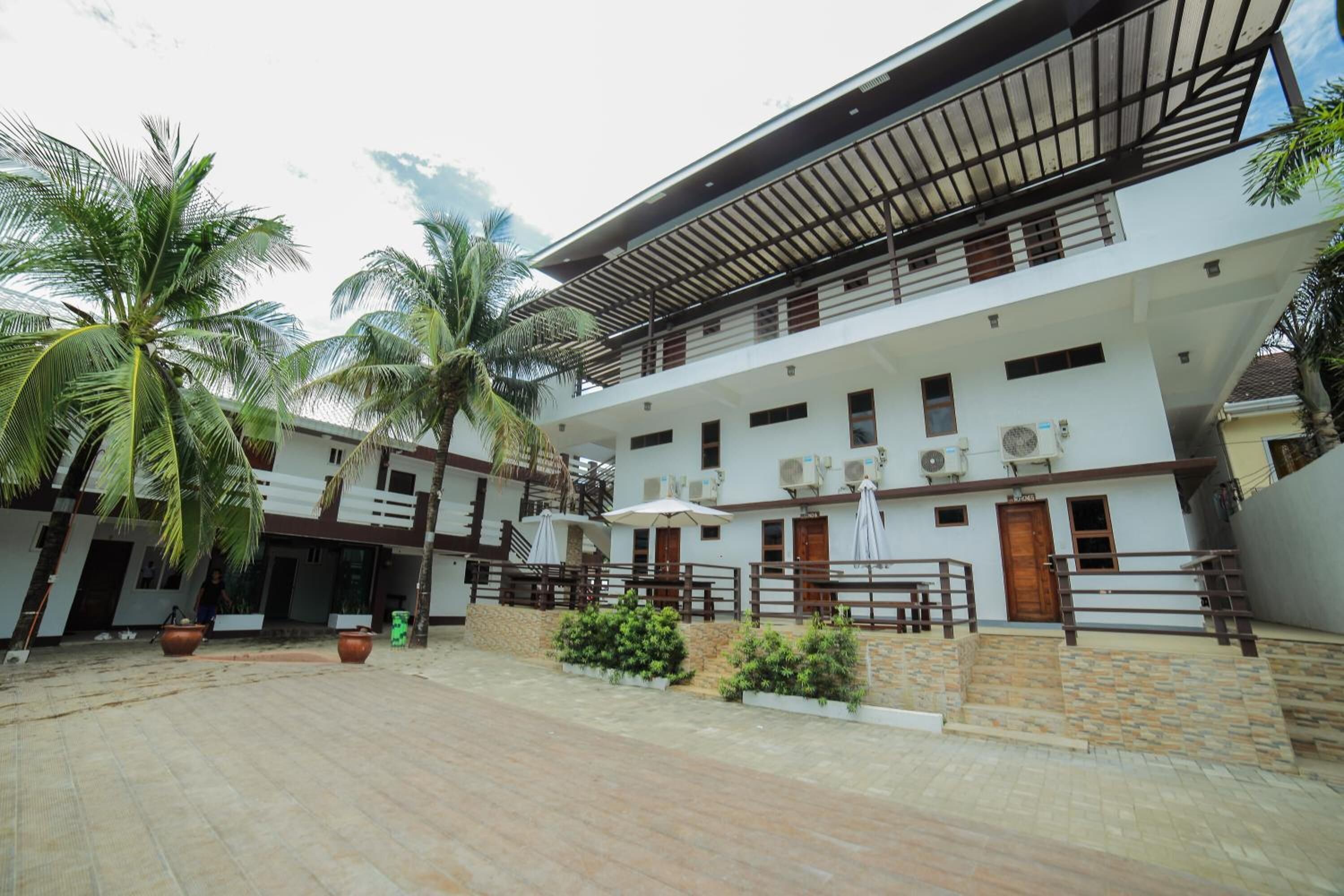 A modern two-story building with a courtyard, featuring a covered outdoor seating area, palm trees, and a stone pathway.