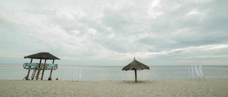 A beach with two thatched umbrellas and a wooden structure with a ladder.