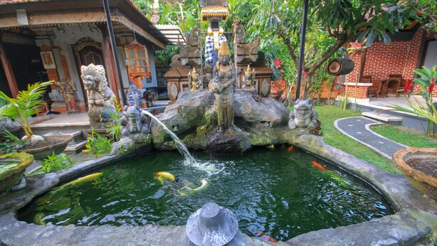 A traditional stone fountain with statues, surrounded by lush greenery and a brick building in the background. - Pramewari Homestay (Ubud)