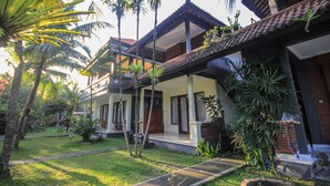 A two-story house with a covered porch, surrounded by a well-maintained garden with palm trees and other tropical plants. - Pramewari Homestay (Ubud)