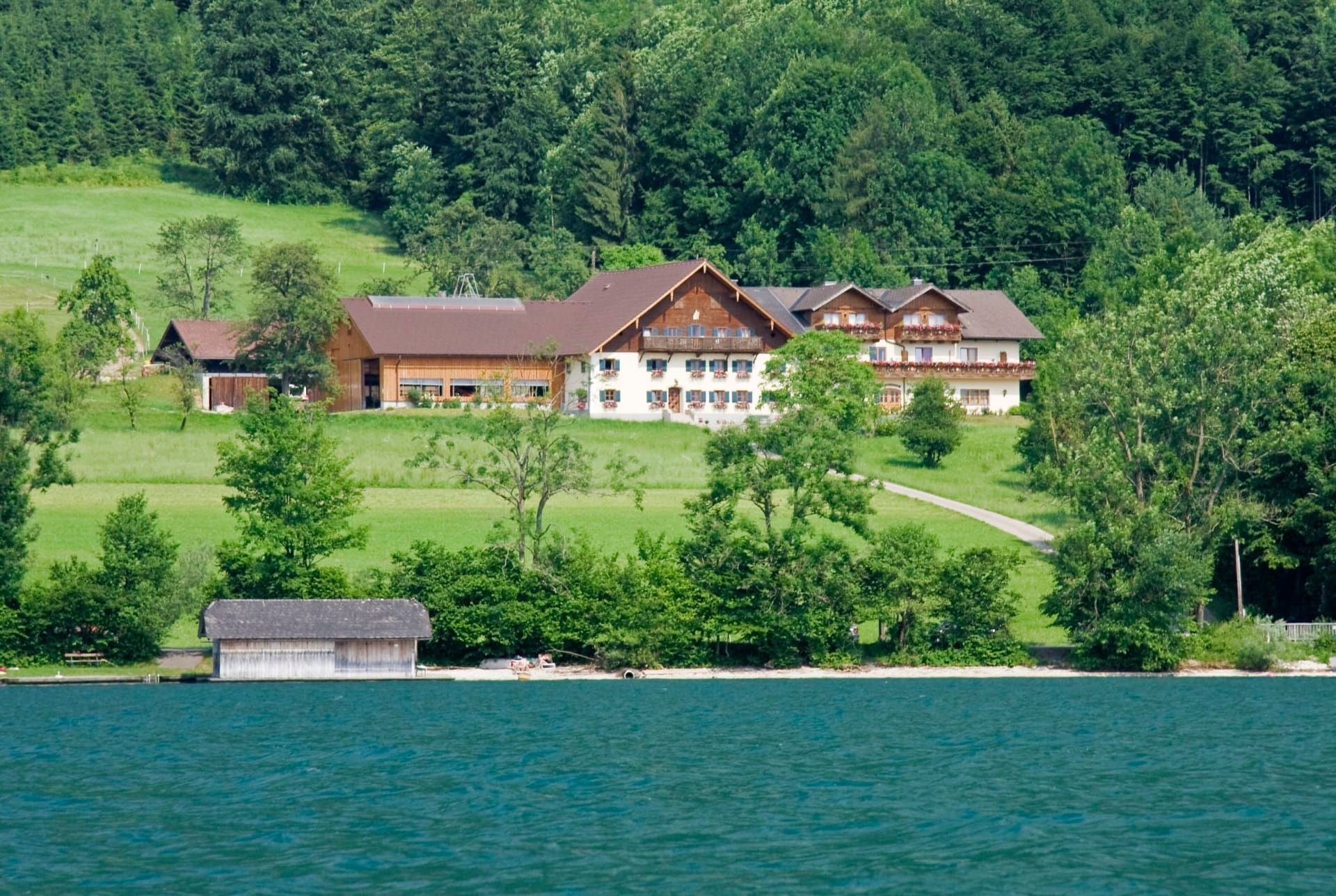 Un grand bâtiment à étages, doté d’une section centrale imposante et de deux ailes plus petites, entouré d’une végétation luxuriante et bordé d’un plan d’eau.
