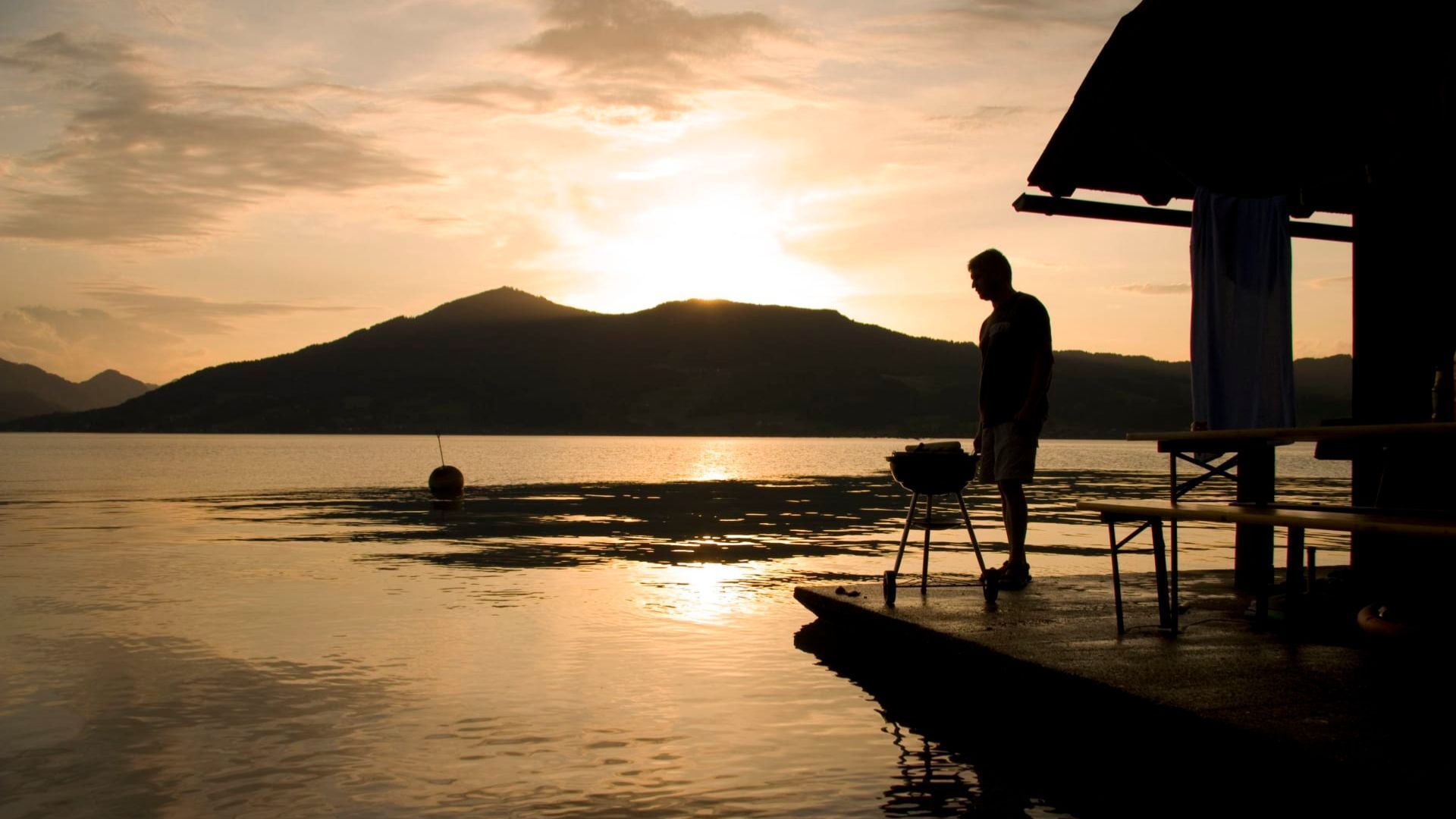 Une personne se tient près d’un quai, à côté d’un bateau, et contemple un lac paisible, avec des montagnes qui se dessinent à l’horizon, au moment du coucher du soleil.