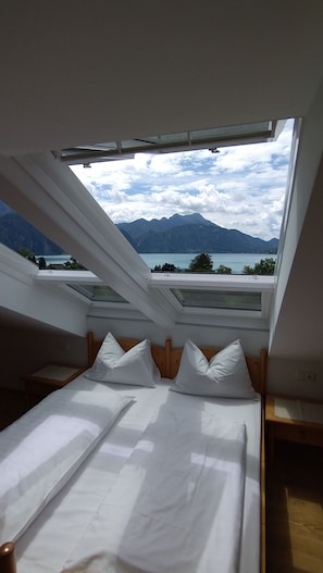 A bedroom with a skylight, a bed with white bedding, and a view of mountains and a lake.