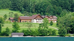 A large, multi-story building with a prominent central section and two smaller wings, surrounded by lush greenery and a body of water. - Apartment Brennerin - Haslbauer - family Resch (Haslach)