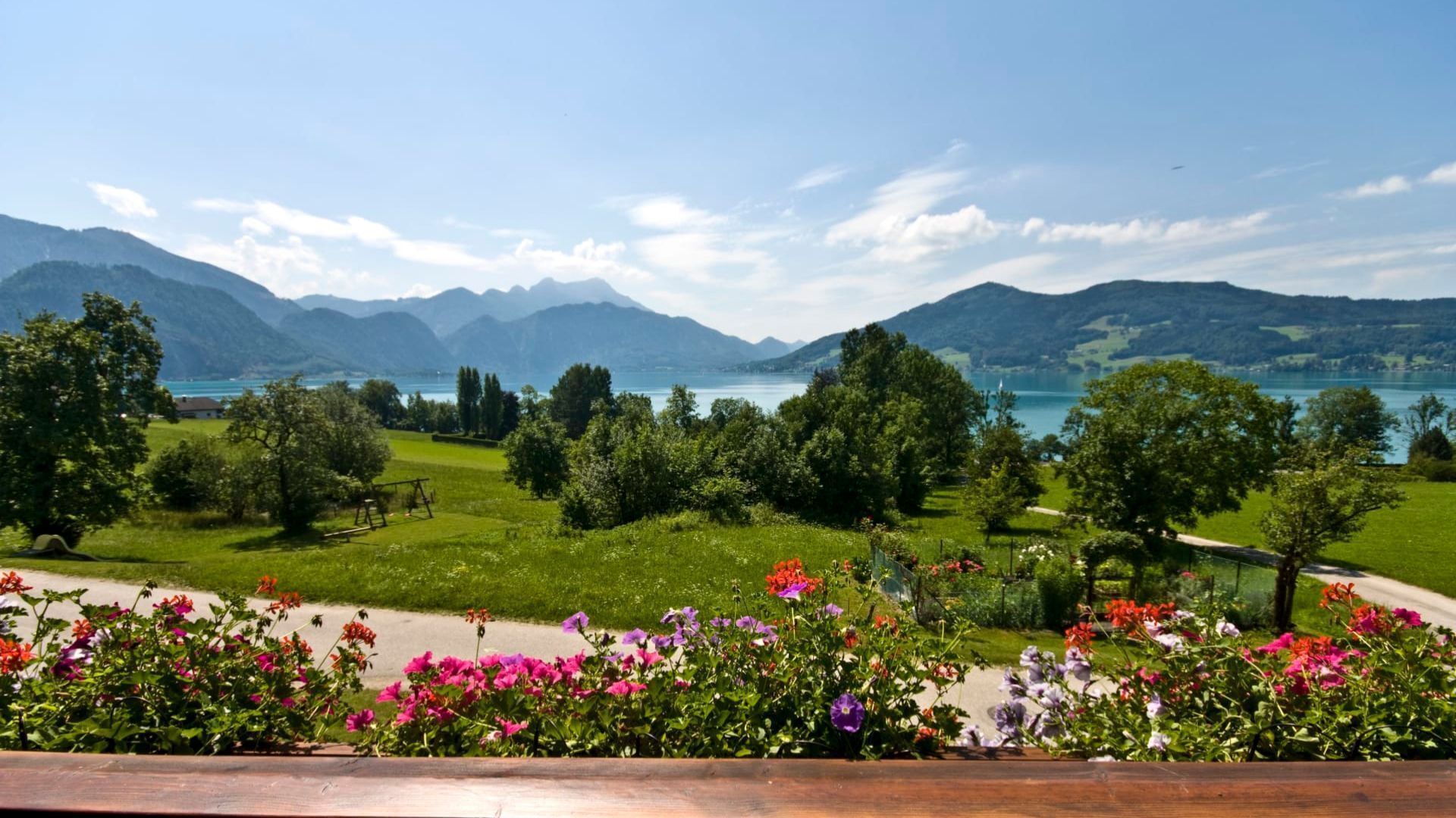 Vista panorâmica de um lago rodeado por montanhas, com uma trilha e canteiros de flores ao primeiro plano.