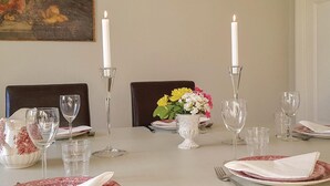Una mesa de comedor dispuesta con platos blancos, vasos transparentes y una jarrón blanco. Dos velas altas están colocadas en candelabros de vidrio. Un jarrón con flores amarillas y blancas se encuentra sobre la mesa.