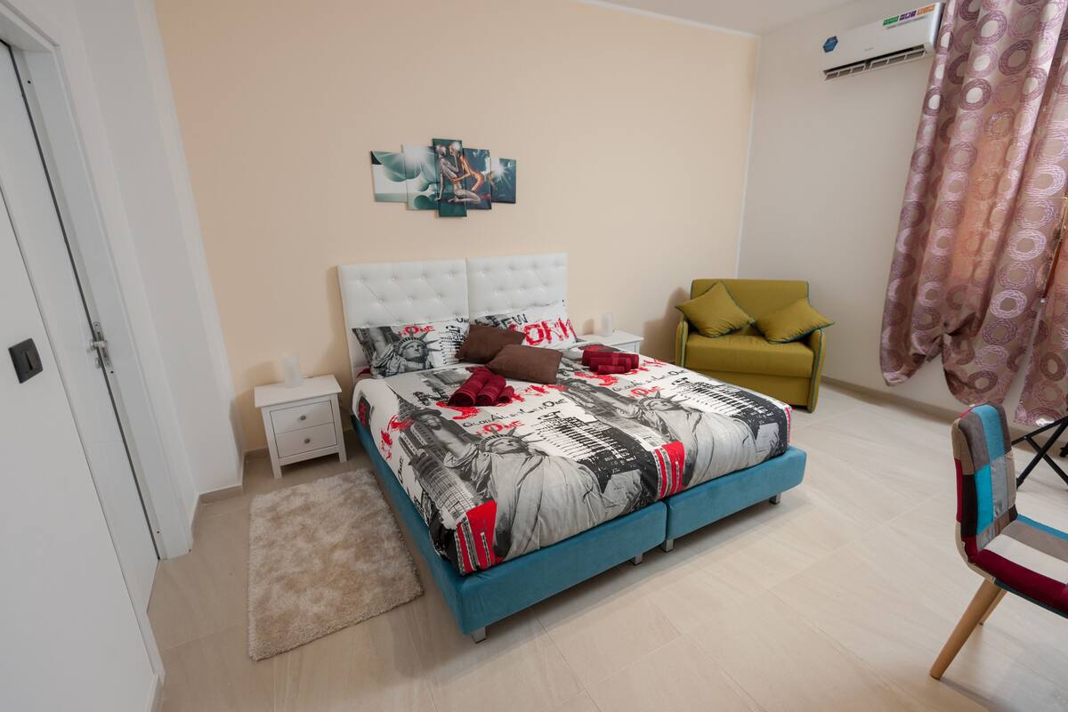 A bedroom with a bed, a nightstand, a chair, and a wall-mounted air conditioner.