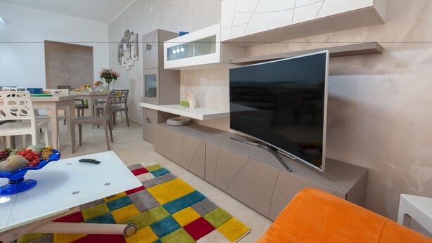 Una sala de estar moderna con un televisor de pantalla plana, una zona de comedor con una mesa y sillas, y una alfombra colorida.