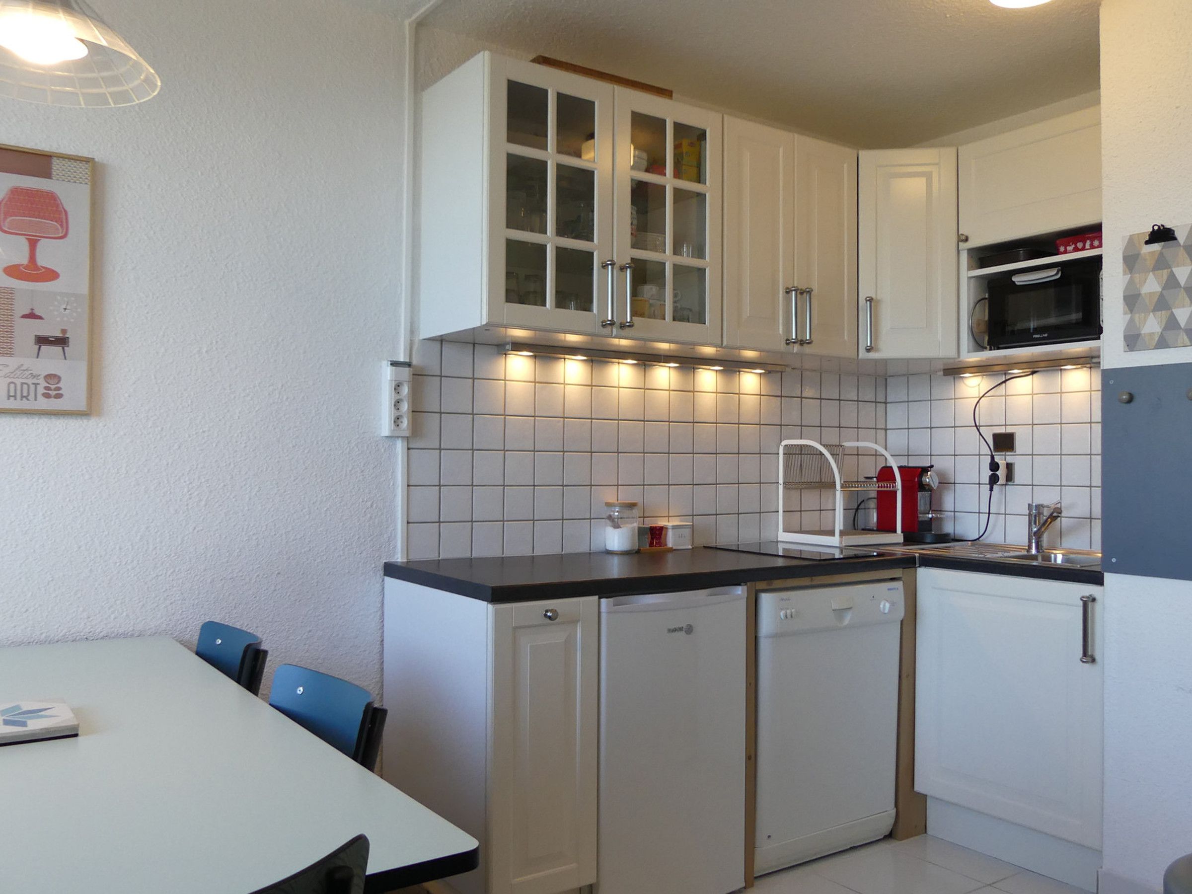 A compact kitchen with white cabinets, a microwave, and a dishwasher. There is a dining table with chairs and a wall-mounted light fixture.