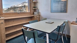 Une pièce chaleureuse avec une grande fenêtre donnant sur une montagne enneigée, un banc en bois, une table avec des chaises et une œuvre d’art abstraite encadrée accrochée au mur.