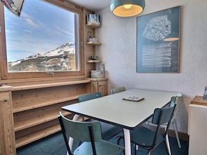 A cozy room with a large window overlooking a snowy mountain, a wooden bench, a table with chairs, and a framed abstract art piece on the wall.