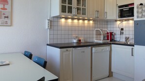 Une cuisine compacte avec des meubles blancs, un four à micro-ondes et un lave-vaisselle. On y trouve une table à manger avec des chaises et un luminaire fixé au mur.