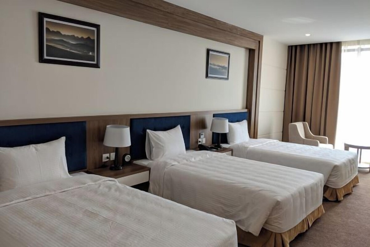 A hotel room with two beds, a wooden headboard, bedside lamps, a chair, and a view of the outdoors.