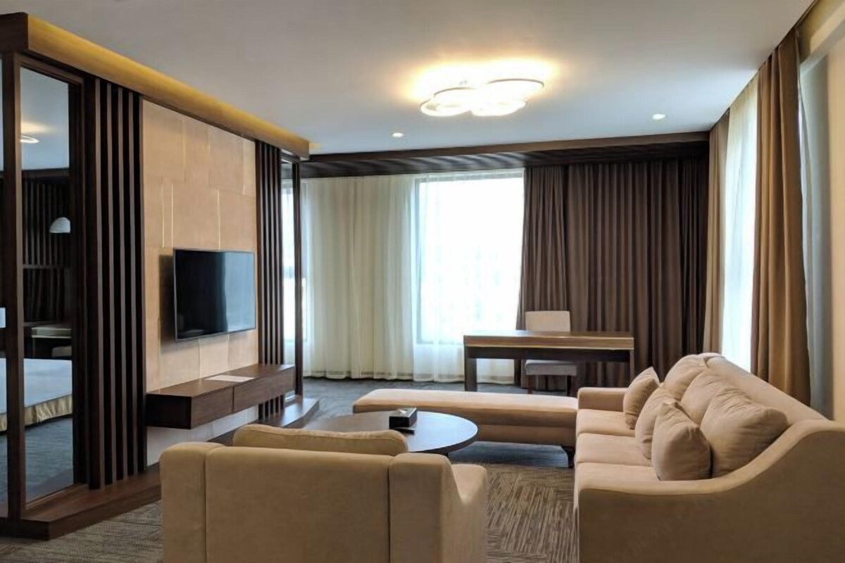 Ein modernes Hotelzimmer mit einer Couch, einem Couchtisch, einem an der Wand montierten Fernseher, einem Esstisch und einem großen Fenster mit Vorhängen.