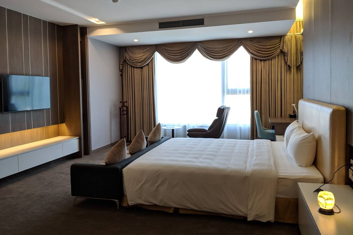 Una habitación de hotel moderna con una cama grande, un televisor de pantalla plana en la pared, una zona de estar con una mesa y sillas, y vistas al exterior a través de amplios ventanales.