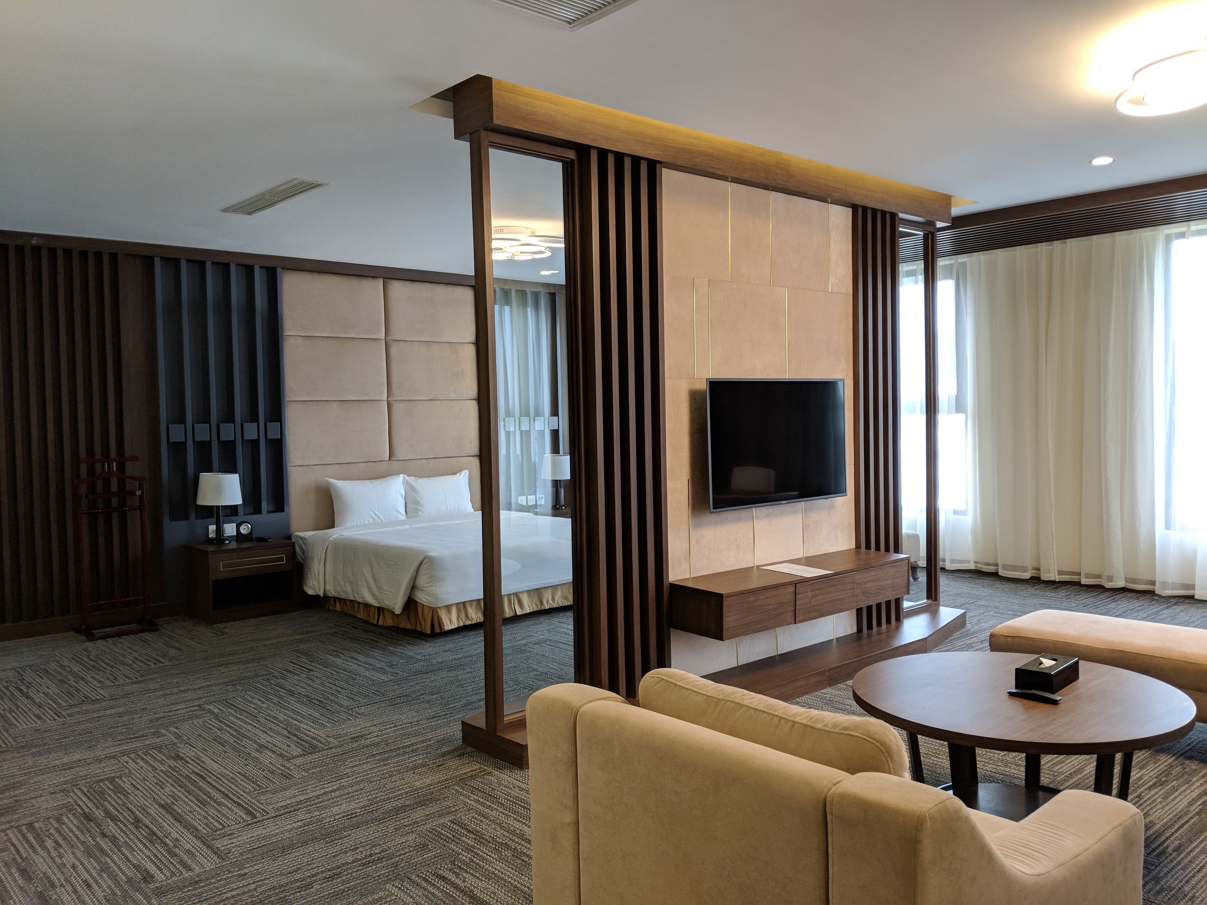 Ein modernes Hotelzimmer mit einem großen Bett, einem flachbildfernseher an der Wand, einer Sitzecke mit rundem Tisch und einem großen Fenster mit Vorhängen.