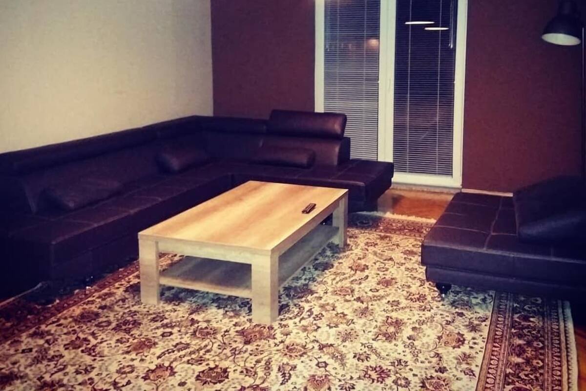 Ein Wohnzimmer mit einem dunkelbraunen Sofa, einem Holztisch und einem gemusterten Teppich. Zwei Pendelleuchten hängen von der Decke.
