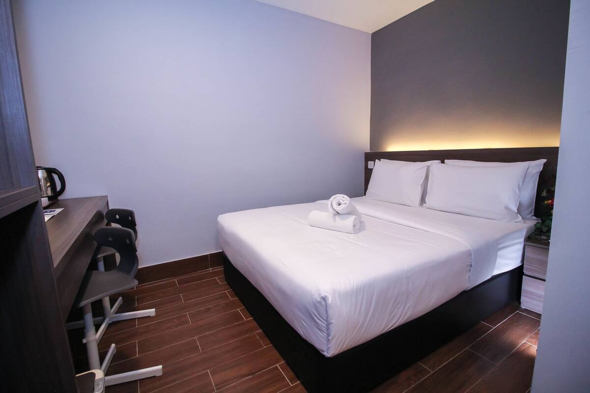 A hotel room with a neatly made bed, a desk with chairs, and a wall-mounted TV.