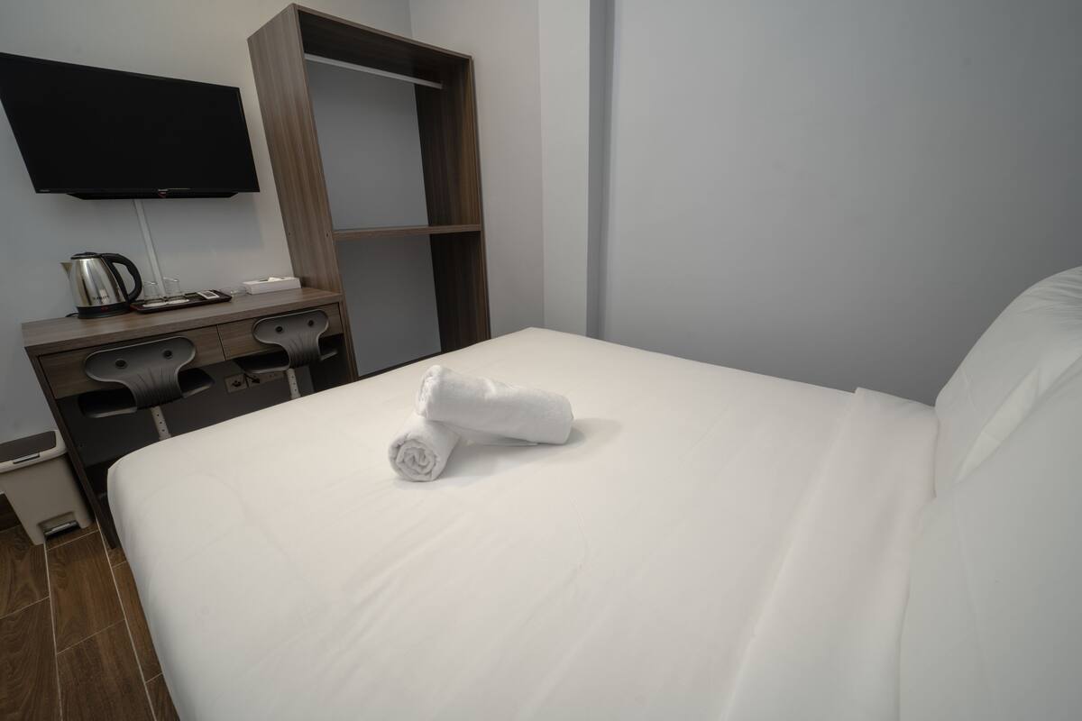A neatly made bed with white linens and a rolled towel, a wooden dresser with a flat-screen TV above it, and a small trash bin beside the dresser.