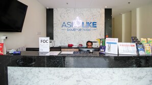 Un comptoir d’accueil en marbre, un grand écran de télévision et un panneau lumineux néon indiquant « ASIA LIKE, hôtel-boutique ».
