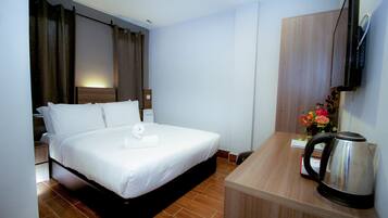 A hotel room with a bed, a desk, a kettle, and a TV mounted on the wall.