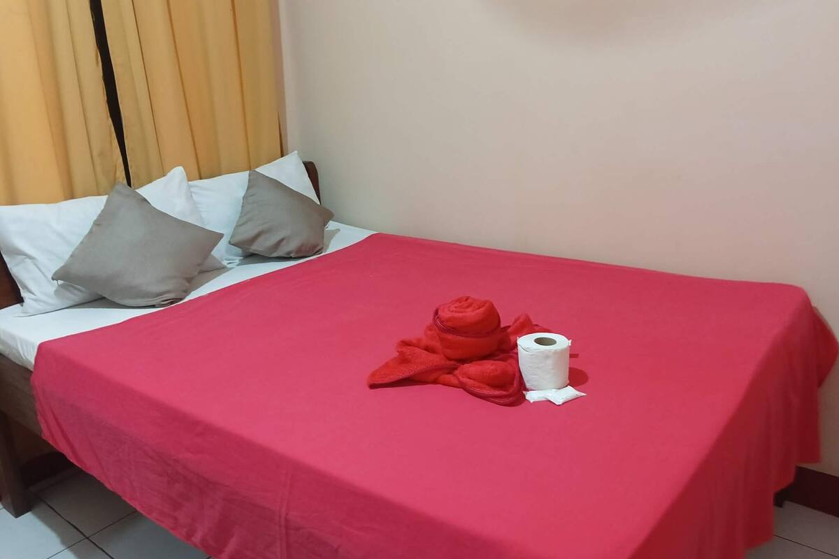 A neatly made bed with a red bedspread, pillows, and a rolled-up towel.