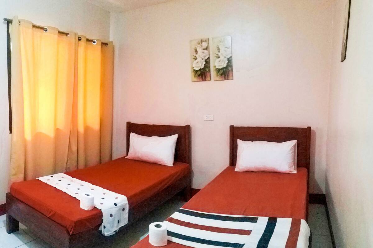 A room with two beds, each with a pillow and a blanket. There is a curtain on one side and a wall with two flower paintings.
