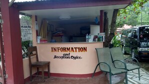 A small reception area with a sign reading "INFORMATION and Reception Office." There are two chairs in front of the counter, and a van is parked outside. - Port Barton DIY (San Vicente)
