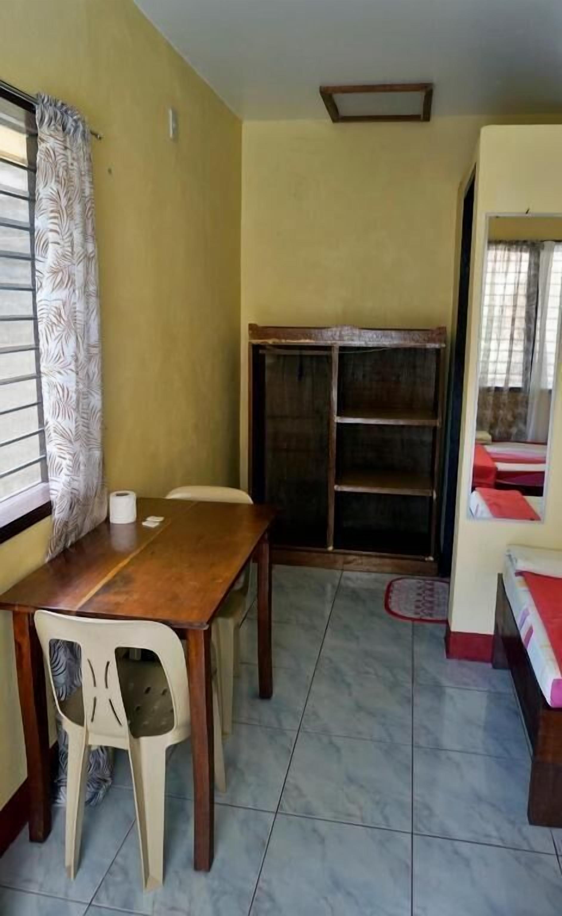 A room with a wooden table, two chairs, a window with patterned curtains, a shelf unit, and a bed with red bedding.