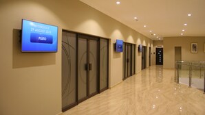 A hallway with a flat-screen TV displaying an event date and name, glass doors, and a polished floor. - Crossroads Hotel Blantyre (Blantyre)