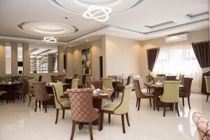 Un comedor de hotel con varias mesas y sillas, iluminación empotrada en el techo y un amplio ventanal con cortinas.