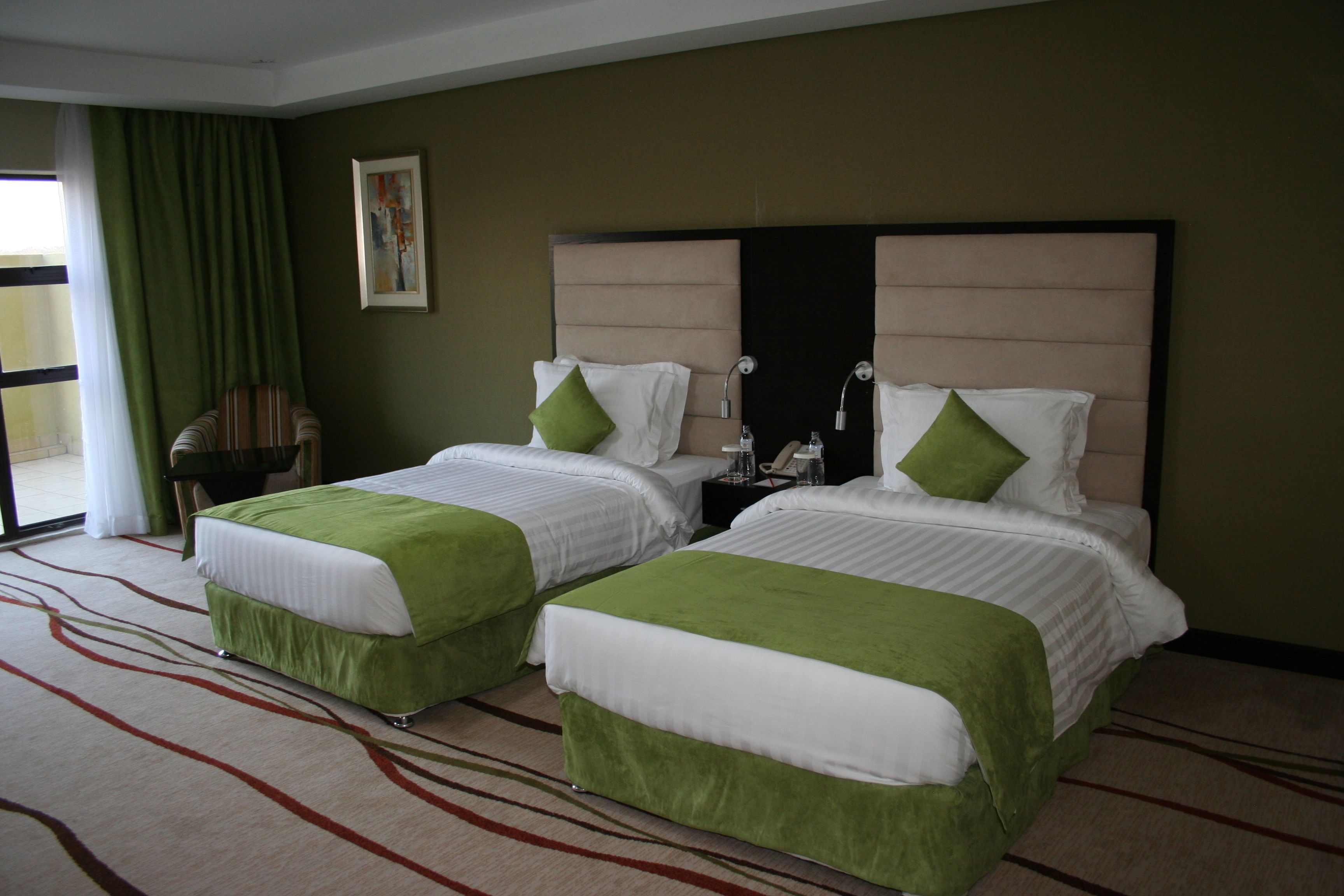 Habitación de hotel con dos camas, cubrecamas verdes, un ventanal con cortinas y un cuadro en la pared, además de una alfombra estampada.
