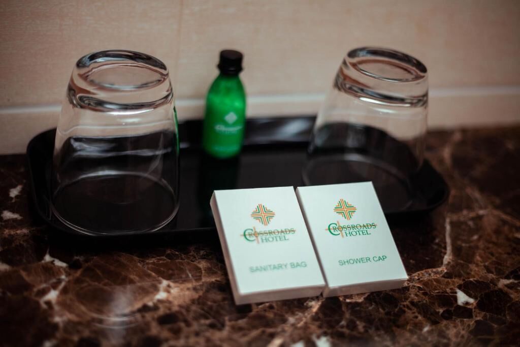 Una bandeja con dos vasos vacíos, una botella verde y dos cajas con la etiqueta 'Crossroads Hotel', indicando 'Bolsa Sanitaria' y 'Gorro de Ducha'.