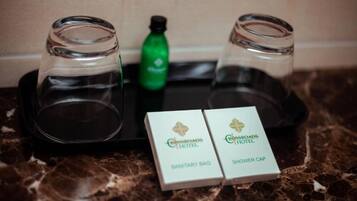 Una bandeja con dos vasos vacíos, una botella verde y dos cajas con la etiqueta 'Crossroads Hotel', indicando 'Bolsa Sanitaria' y 'Gorro de Ducha'.