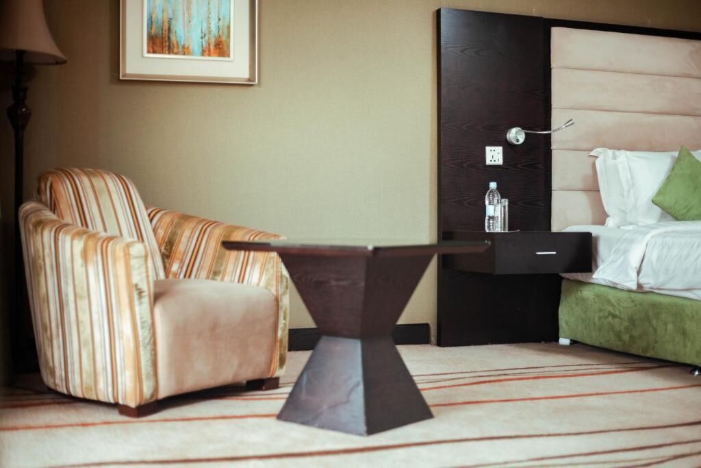 A hotel room with a striped armchair, a bedside table, a bed with green pillows, and a framed picture on the wall.