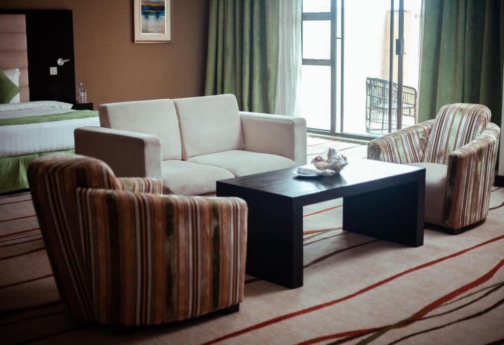Una habitación de hotel con una cama, un sofá, dos sillones a rayas, una mesa de centro y vistas al exterior a través de una ventana.