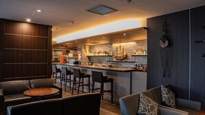 A modern bar area with a wooden bar counter, multiple stools, and a seating area with leather chairs and a round table. - Roygent Parks Hanoi (Hanoi)