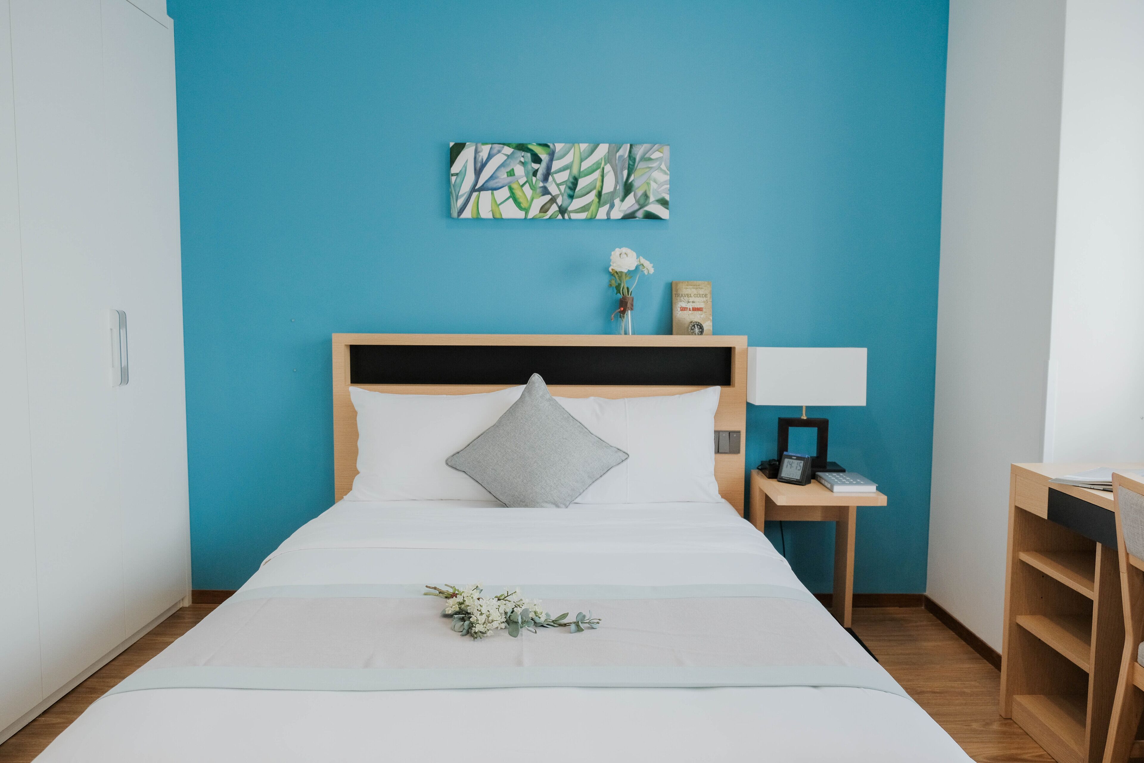 A neatly made bed with a white comforter and a single gray pillow, a small bedside table with a lamp, a framed picture on the wall, and a small vase with a white flower.