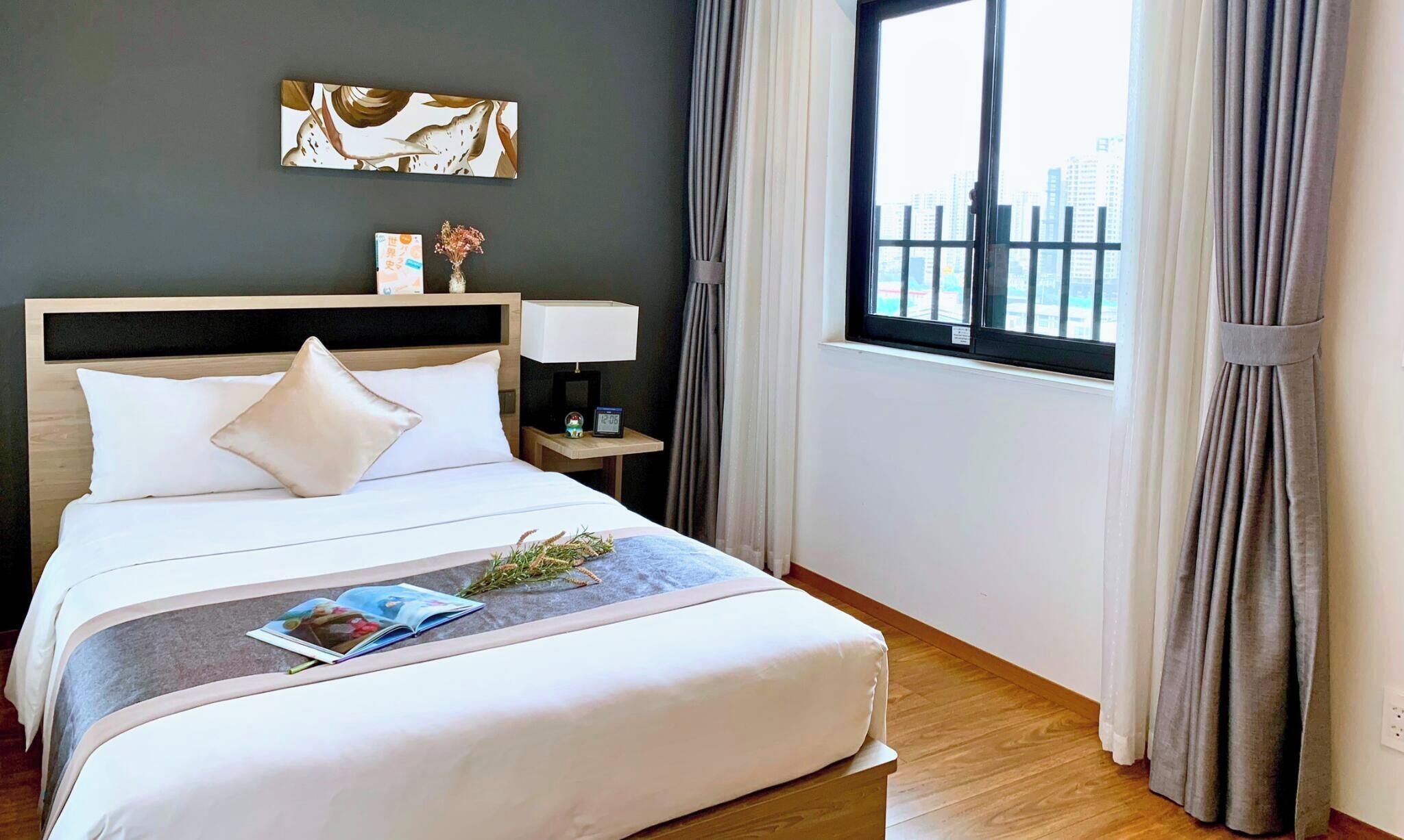 A hotel room with a bed, a nightstand, a lamp, a clock, a window with curtains, and a city view.